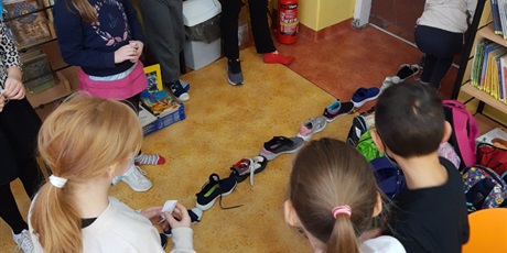 Powiększ grafikę: Uczniowie stoją z zdjętym jednym butem. Obuwie ustawione w prostej lini w kierunku drzwi.